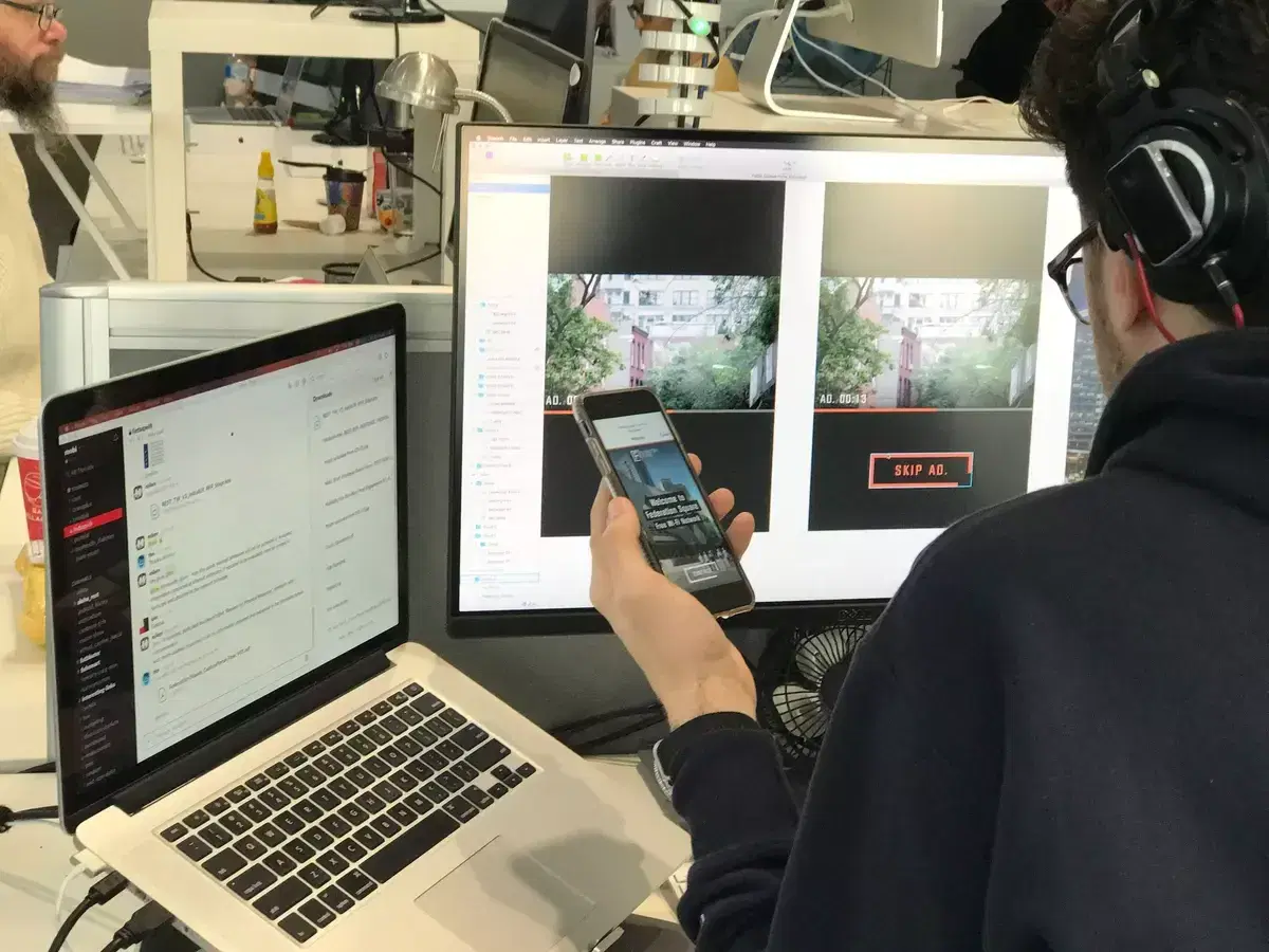 A Mobiddiction developer testing a mobile app on a phone beside design screens on a monitor