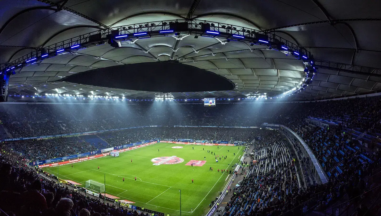 A packed football stadium at night under floodlights, seen from the upper stand