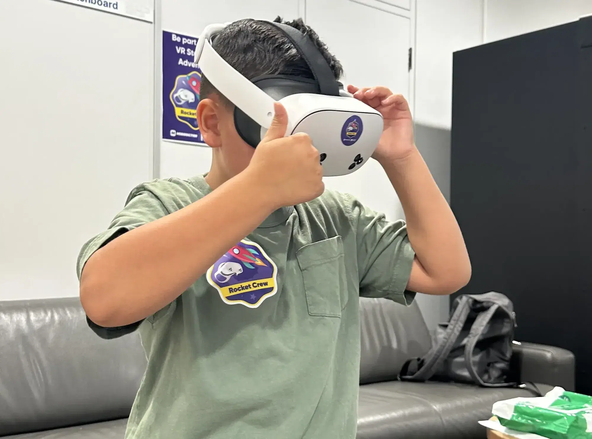 A boy with a Rocket Crew sticker on his shirt puts on a VR headset and gives a thumbs up