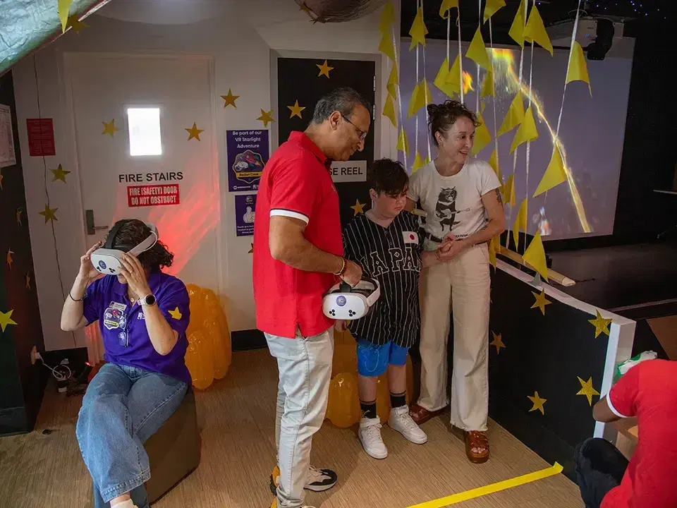 In a Starlight Express Room decorated with stars, a woman tries a VR headset while Mike Vasavada hands a headset to a boy