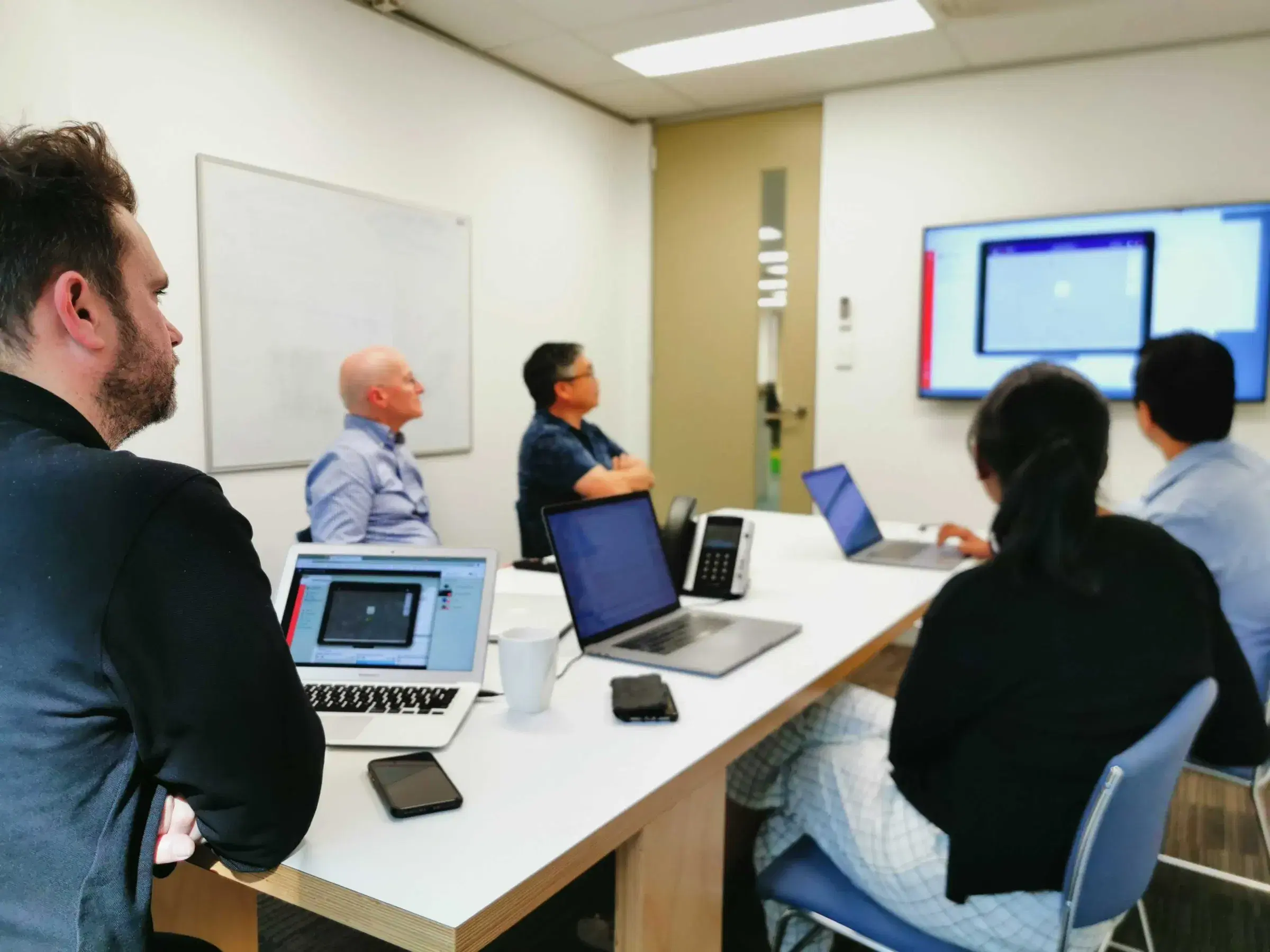 Mobiddiction team members in a meeting room reviewing a program on a wall screen, laptops open on the table