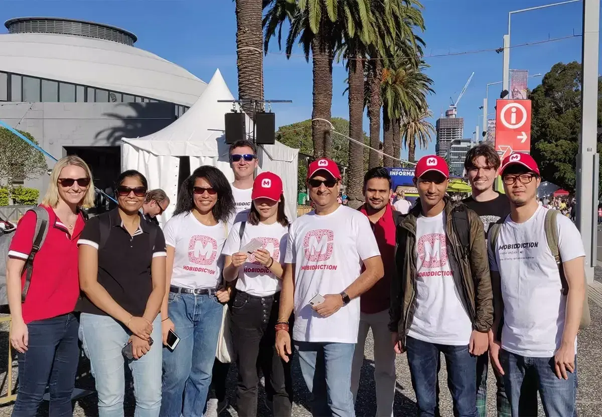 The Mobiddiction team in branded caps and T-shirts, standing together outdoors under palm trees