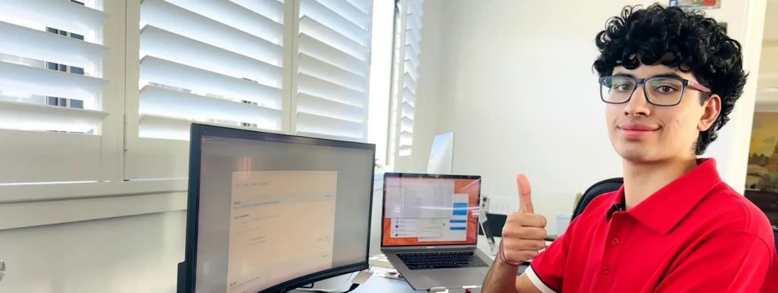 Young man with curly hair and glasses sits at a bright desk, giving a thumbs-up beside two monitors.