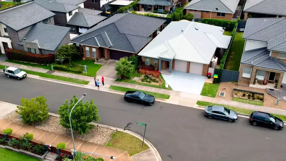 Low drone view of a suburban street, with a white-roofed house, landscaped gardens and parked cars
