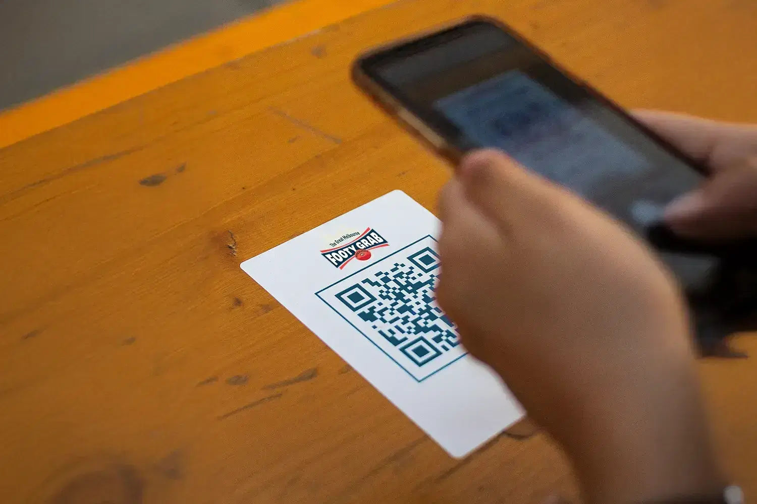 A phone scanning a Great Melbourne Footy Grab QR code card on a wooden table